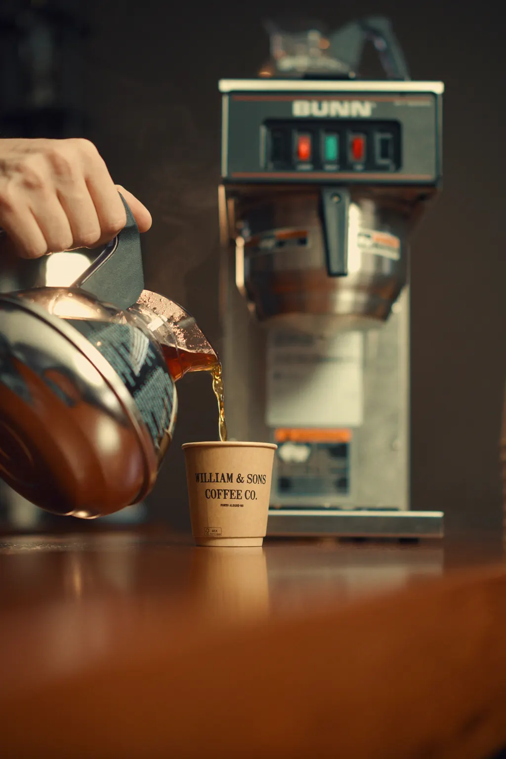 Barista servindo café coado William & Sons Coffee Co. em copo de papel sobre balcão de madeira, com cafeteira profissional Bunn ao fundo
