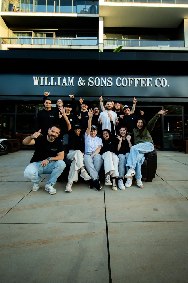 Equipe da William & Sons Coffee Co. reunida e sorrindo em frente à fachada da loja em Porto Alegre, sob o letreiro branco 'William & Sons Coffee Co.'